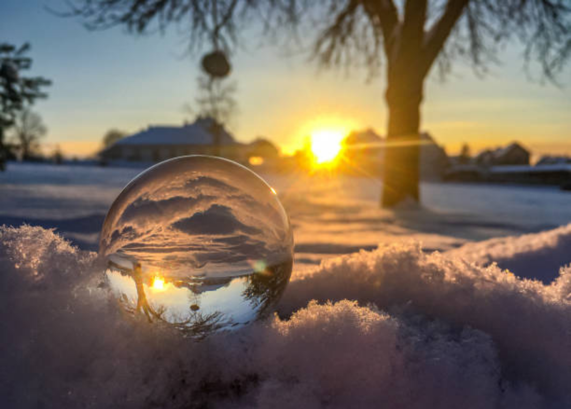 A glass crystal ball sits in the snow in front of a sunset. There are houses in the distance. The background appears as upside down from within the glass ball.