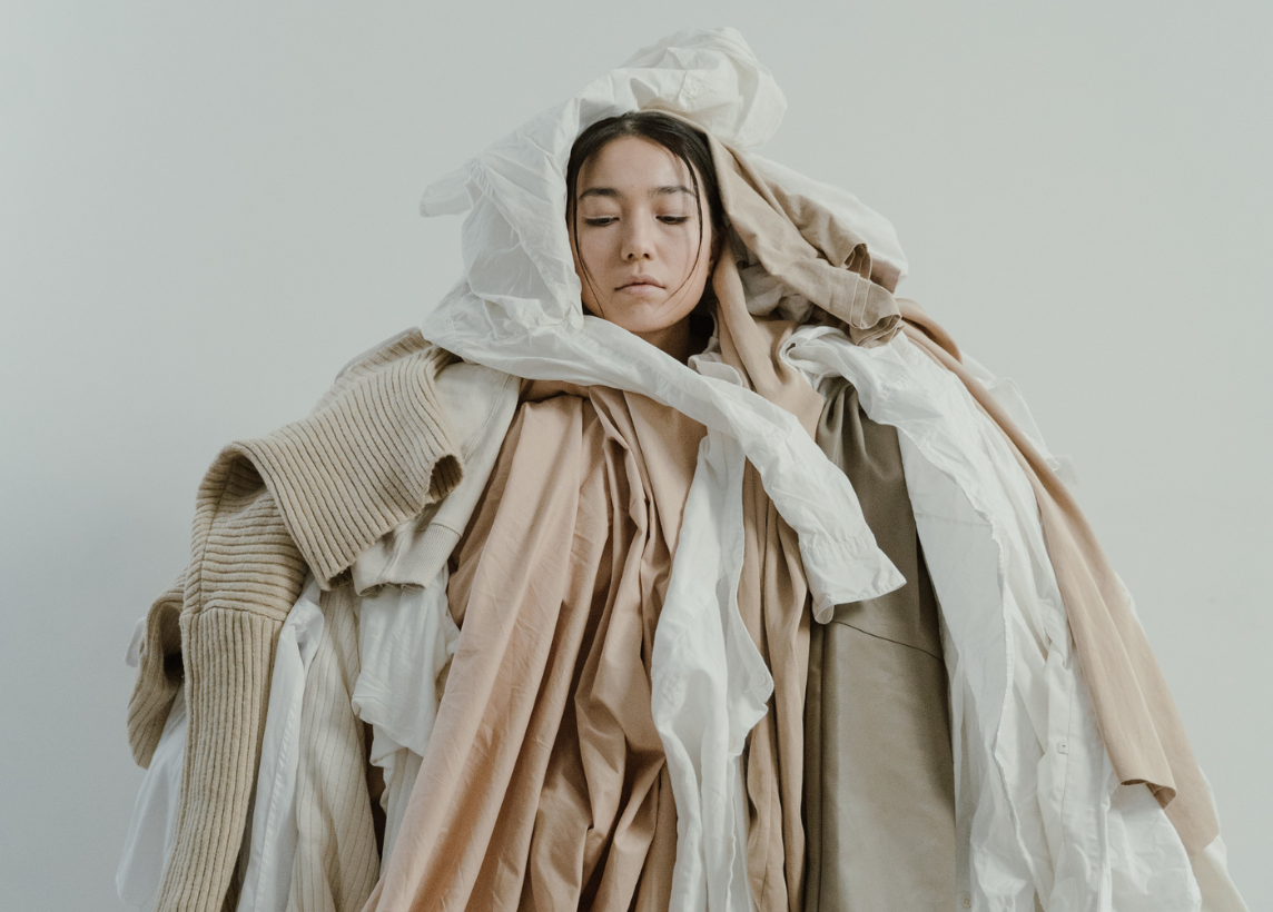 A woman stands with her eyes closed under a pile of neutral colored clothing that has been draped over her head and torso.