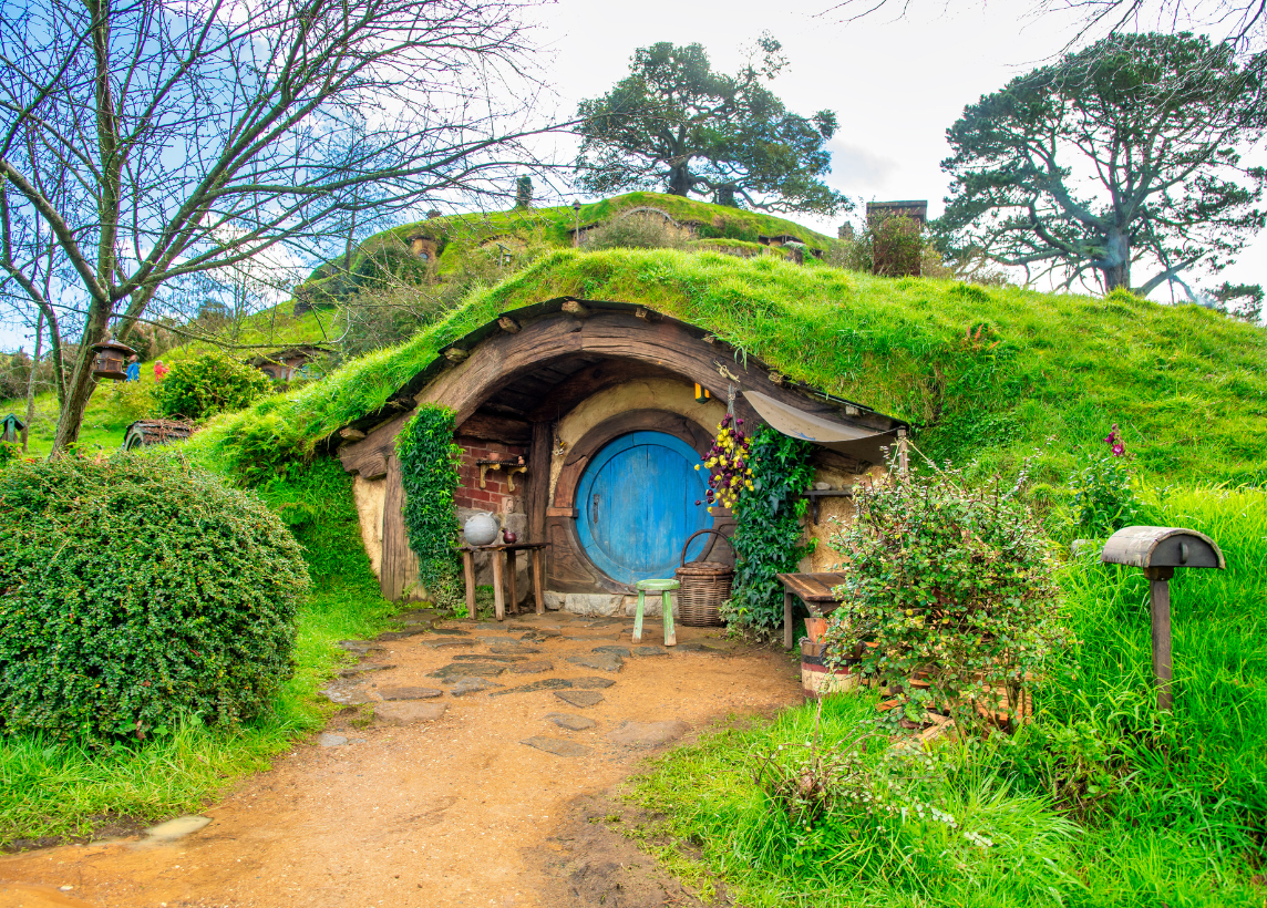 A Hobbit house from the Lord of the Rings movies, built into a green grassy hill with a round door. A dirt trail leads to the door and lush greenery surrounds the entrance over a bright Summer sky.