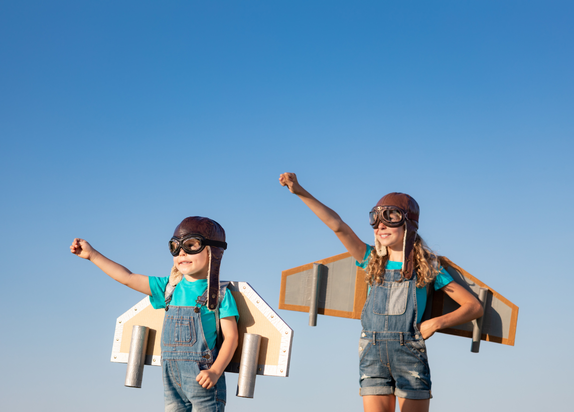 Two young girls pose with one hand pointed at the sky for takeoff! The backdrop is a clear blue sky and they are dressed up with pilot caps and goggles, as well as cardboard wings shaped like a plane.