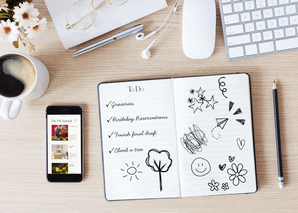 Photo depicts a birds eye view of a desk with a cup of coffee, headphones, keyboard and mouse, and a few flowers sitting near the top. The focus of the image is a to-do list in an open journal in the middle listing groceries, birthday reservations, finishing final draft, and climbing a tree as tasks to complete. The other page is filled with doodles. A phone sitting on the left of the journal is opened to the Virtuosa Society blog page.