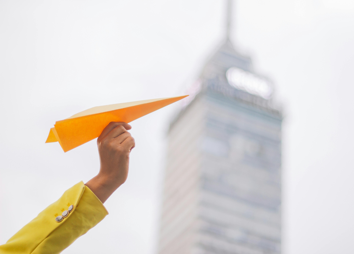 Someone reaches their arm into frame in the lower left of the image. They are wearing a long-sleeve yellow garment and holding a yellow paper airplane. A skyscraper is blurred in the background against an overcast sky.