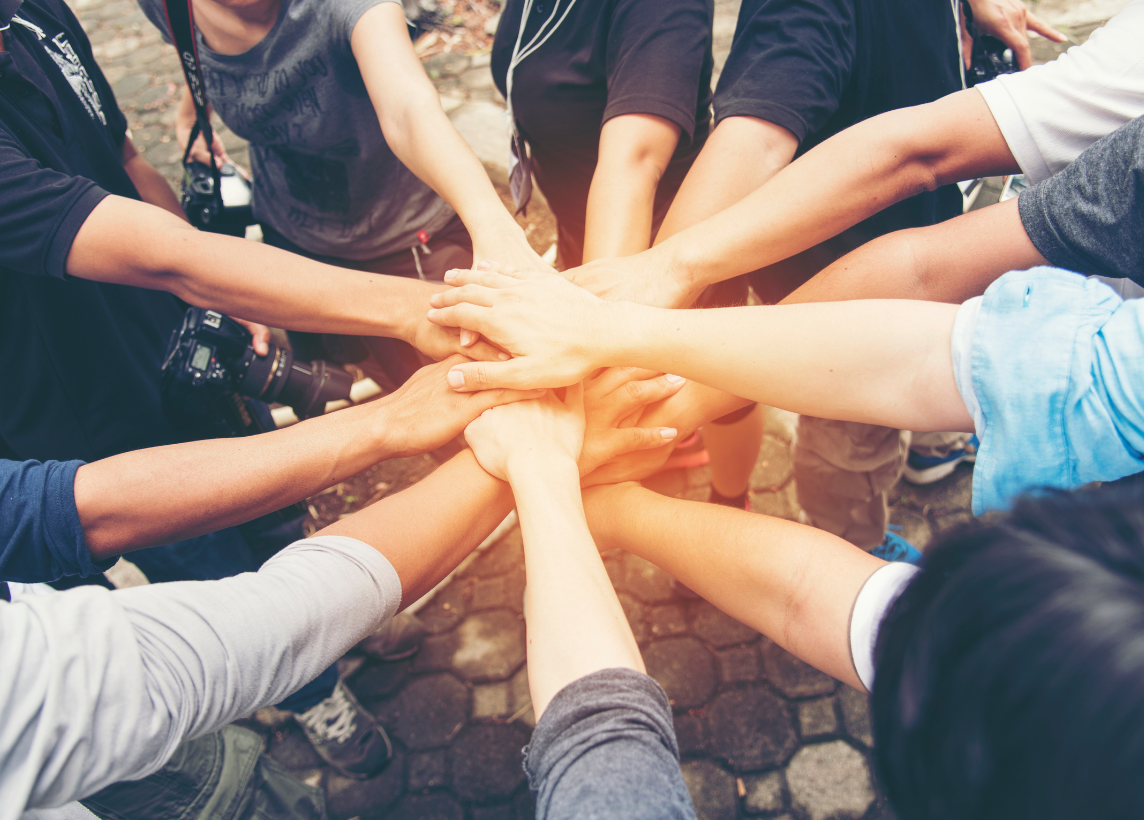 Several people standing in a circle and reaching their hands toward the middle, a common "group handshake" of sorts before beginning a task or project together.