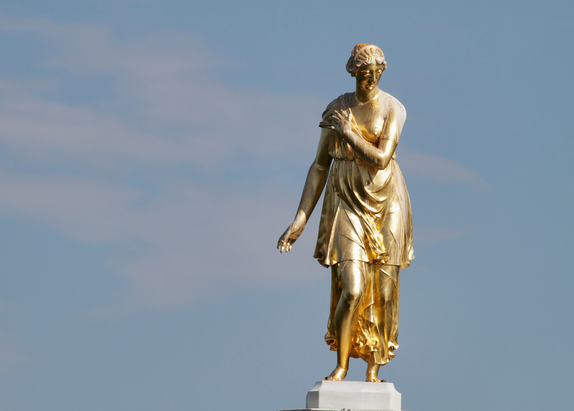 A golden statue worn by time depicts the Roman goddess Diana against a clear blue sky.