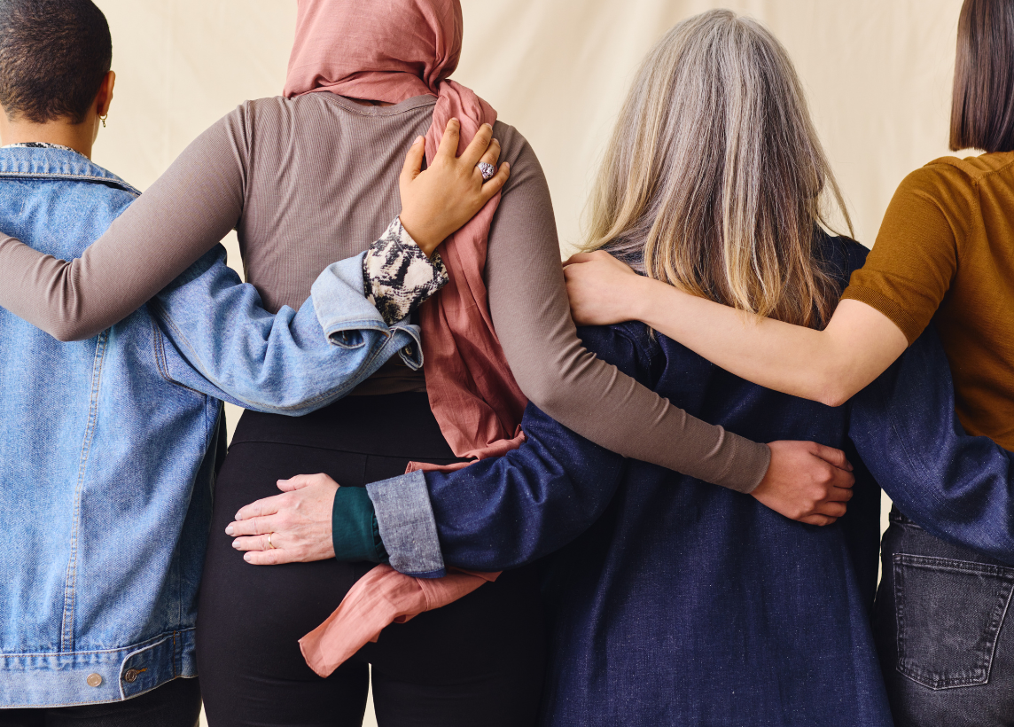 Four women stand with their backs to the camera and arms around each other.